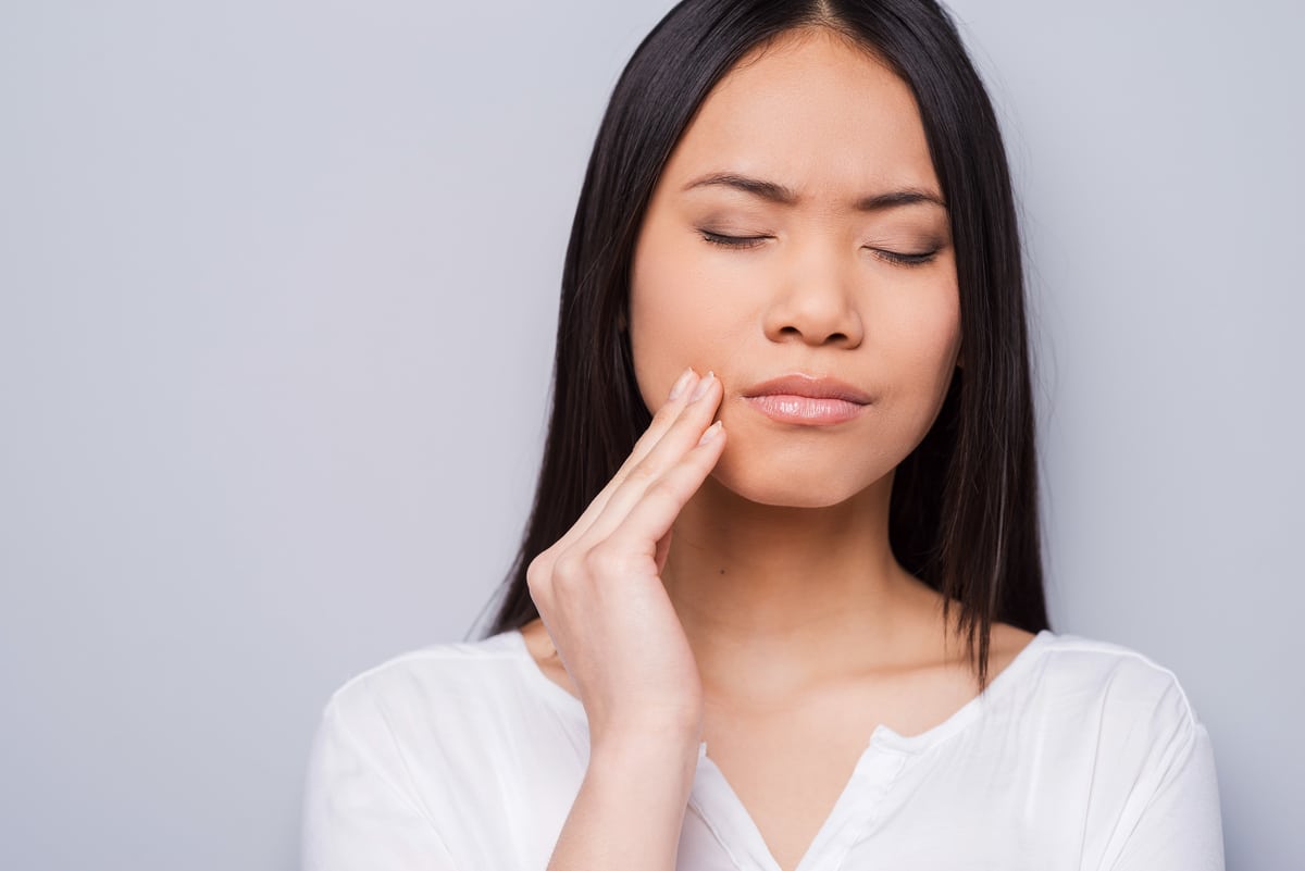 toothache. beautiful young asian women touching her cheek and keeping eyes closed while standing against grey background