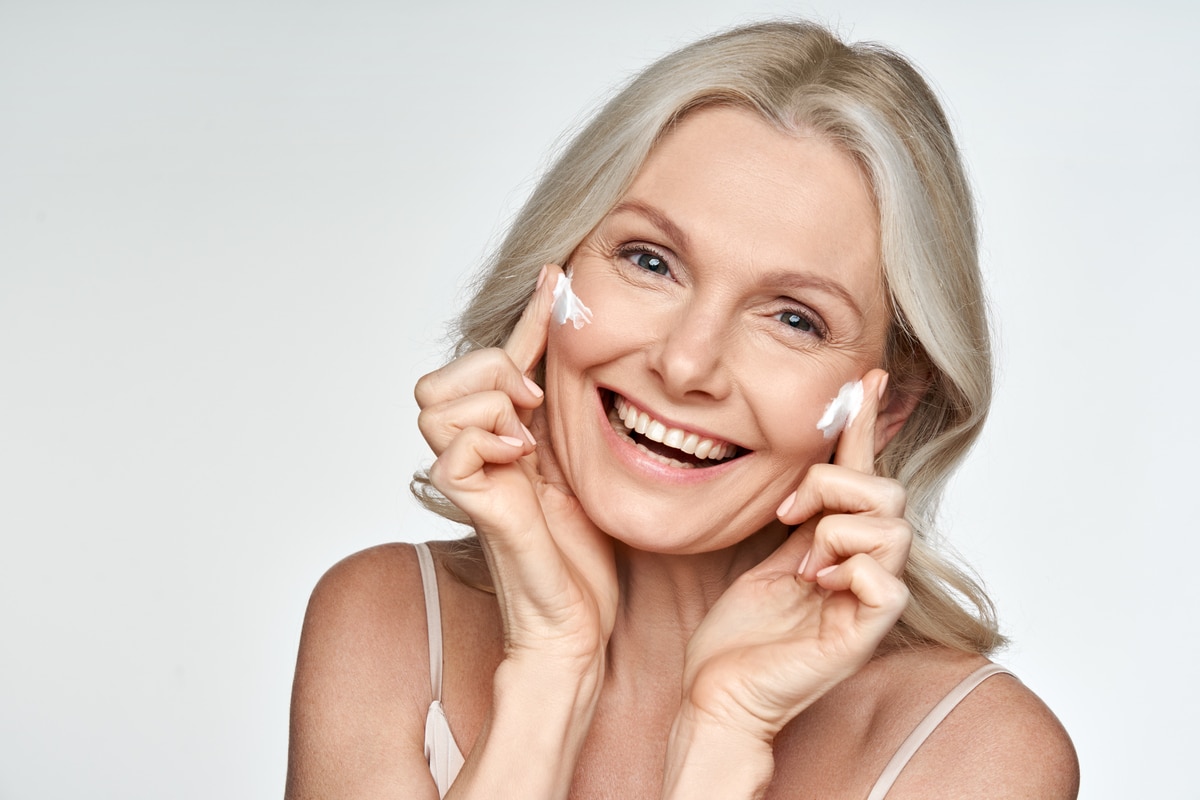 heureuse femme d'âge moyen appliquant de la crème sur la peau du visage isolée sur fond blanc. anti rides