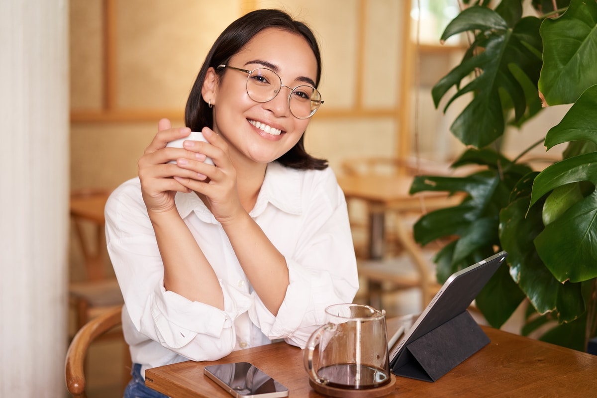 fille asiatique souriante dans des verres, femme travaillant à distance, buvant du café et utilisant une tablette, lunettes de vue tendance