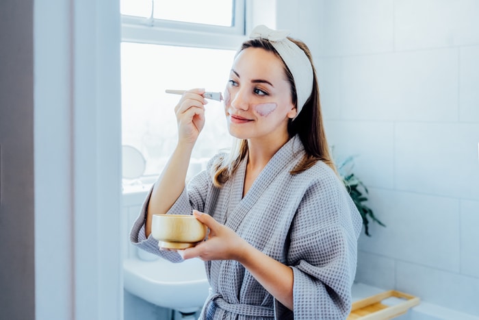 jeune femme en peignoir regardant dans le miroir et appliquant un masque d'argile cosmétique naturel sur son visage dans la salle de bain. procédures cosmétiques pour les soins de la peau à domicile. soins de beauté. mise au point sélective