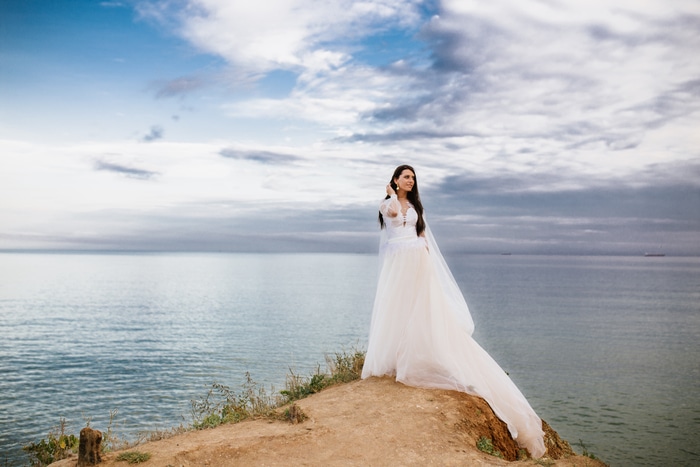 belle mariée romantique en robe blanche posant sur une terrasse avec vue sur la mer et les montagnes en arrière-plan grece