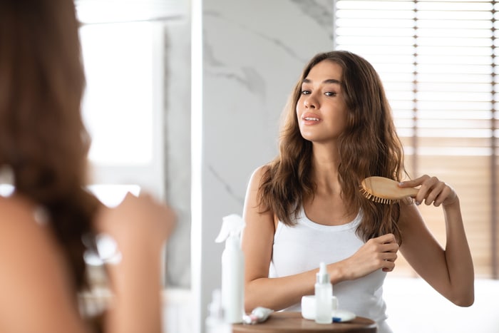jeune femme se brosser les cheveux avec une brosse à cheveux debout dans la salle de bain à l'intérieur