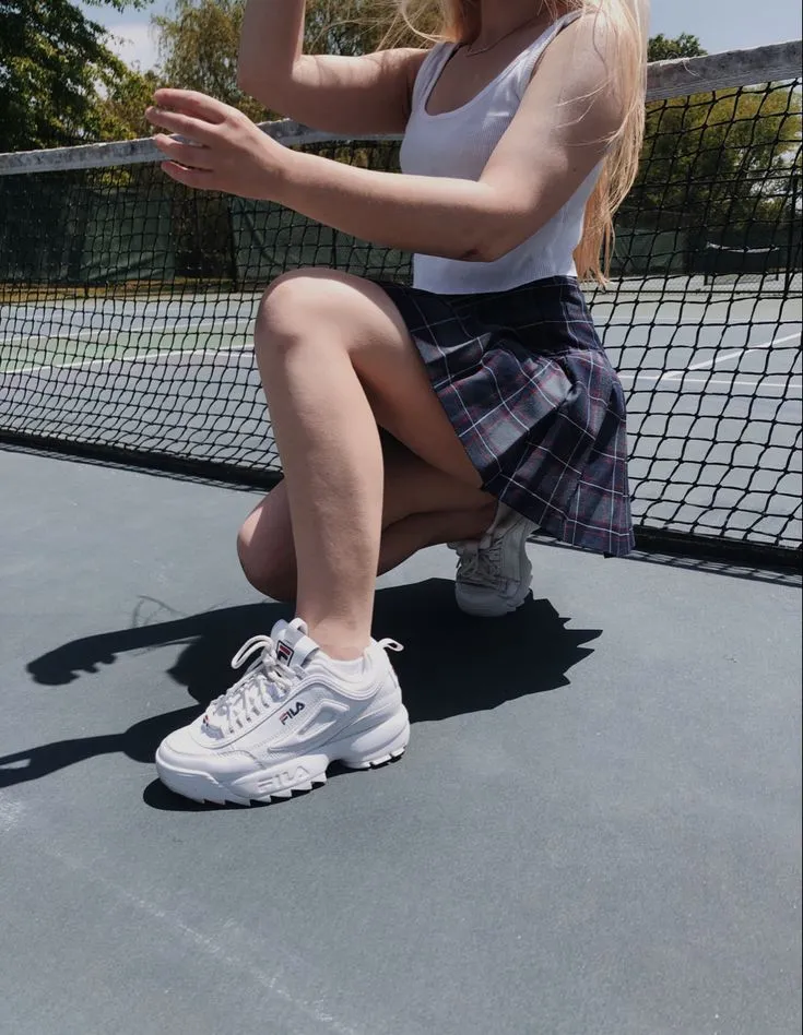 Femme accroupie sur un court de tennis, en jupe à carreaux et Fila Disruptor blanches