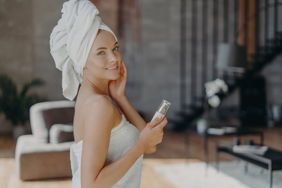 photo intérieure d'une jolie femme européenne avec une peau saine et éclatante, un maquillage minimal, tient une bouteille de lotion pour le corps, dorlote le teint, porte une serviette de bain autour du corps, pose contre l'intérieur domestique