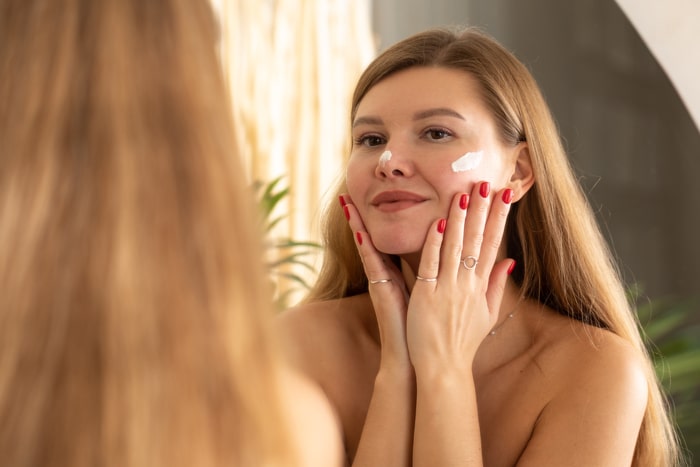 une jolie jeune femme applique une crème hydratante anti-âge sur son visage devant un miroir de salle de bain.