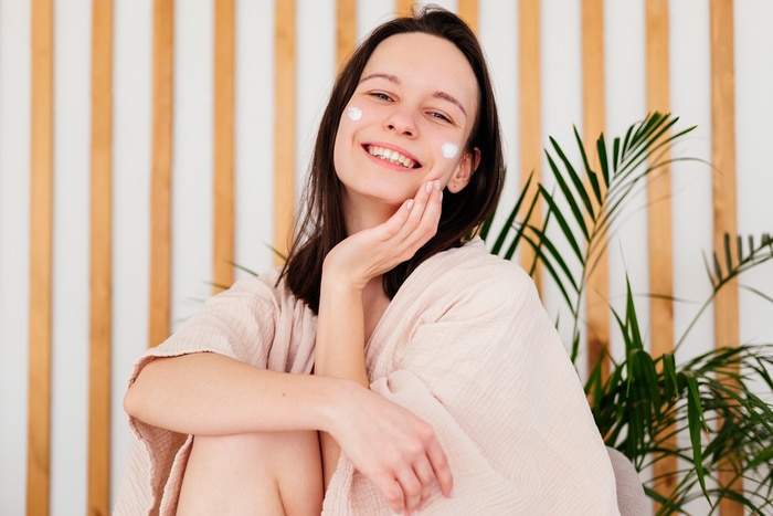 Jeune femme souriante appliquant de la crème sur le visage à la maison en regardant la caméra. Routine beauté du matin. Concept de soins de la peau.