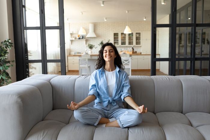 Notion de gestion du stress. Jeune femme paisible en tenue décontractée méditant les yeux fermés sur un canapé à la maison, sur toute la longueur. Femme millénaire calme assise sur un canapé dans une pose de yoga, trouvant l'équilibre intérieur