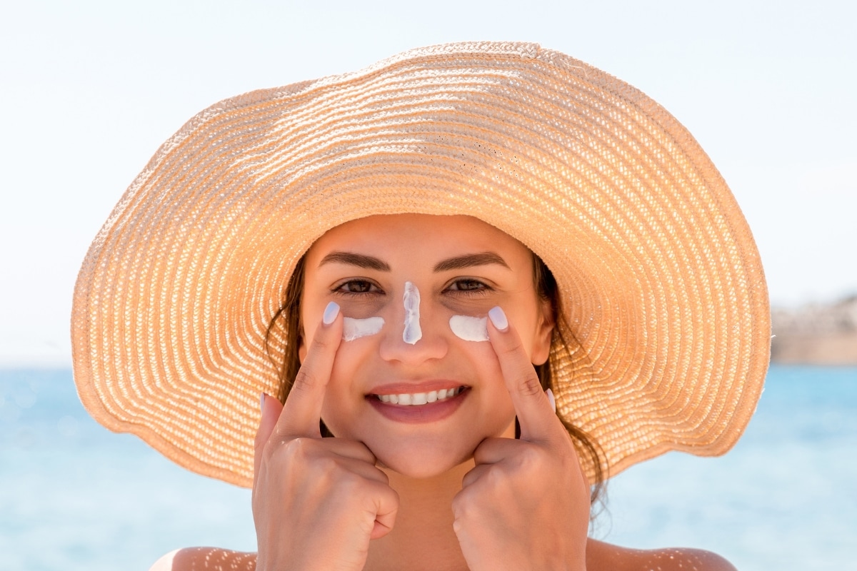 belle femme brune heureuse sur la plage avec un chapeau en paille s'applique de la creme solaire sur le visage