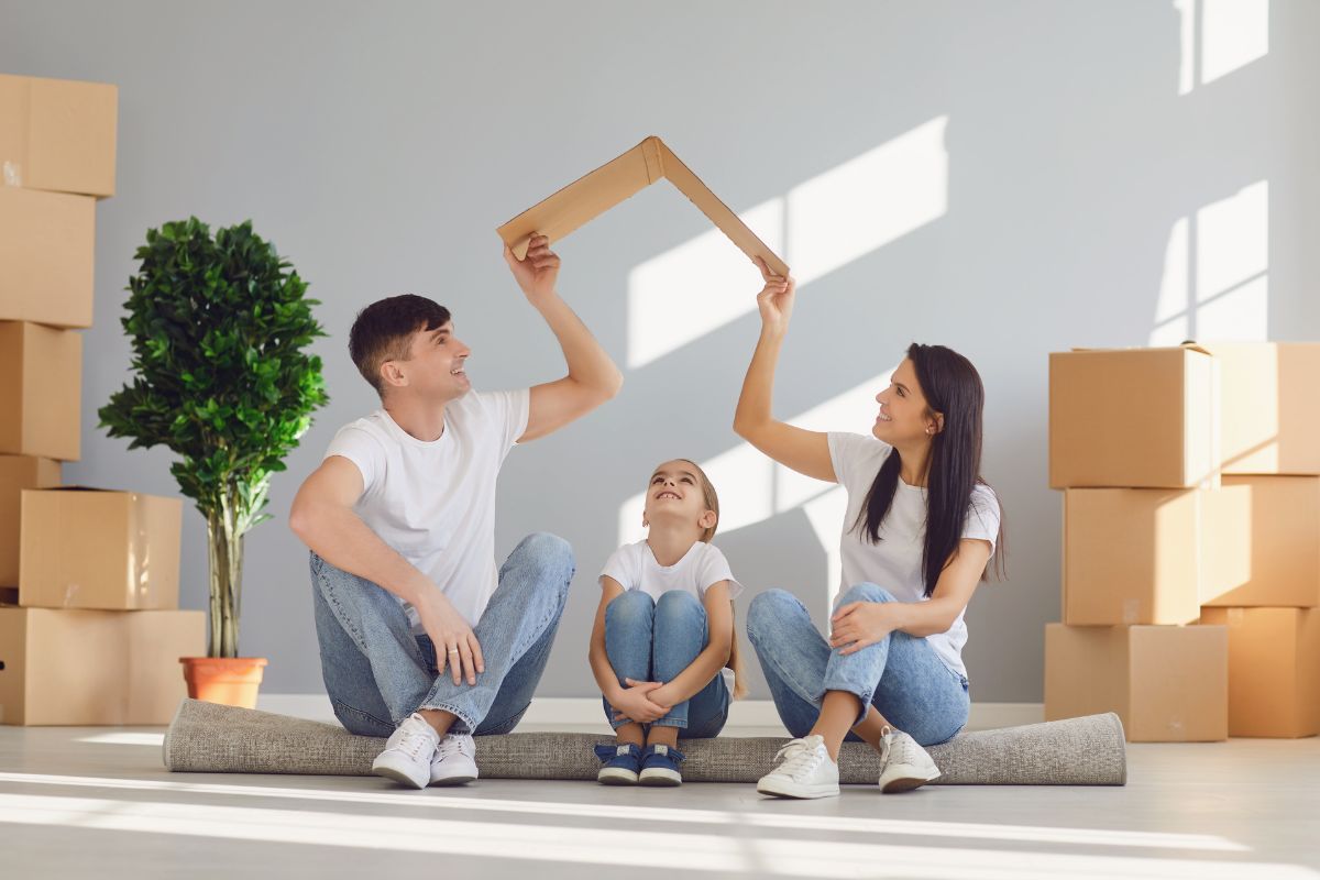 un maman et un papa soulevent un toit en carton au dessus de leur petite fille, symbolisant la protection assurance. Arriere plan de cartons, en plein déménagement