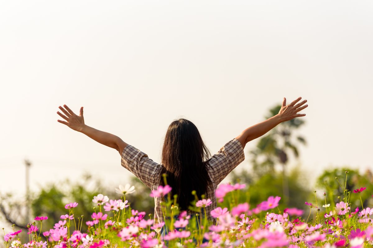 une femme épanouie qui tend les bras au ciel en s'étirant, avec au premier plan une rangée de fleurs roses