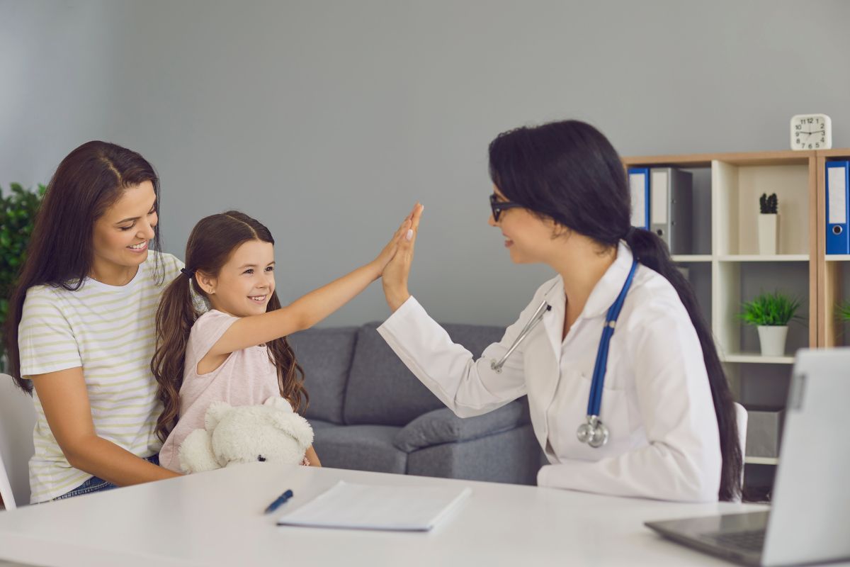 une petite fille tape la main d'une femme docteure en signe de reconnaissance, sur les genoux de sa mère assise, à l'intérieur du cabinet du docteur.