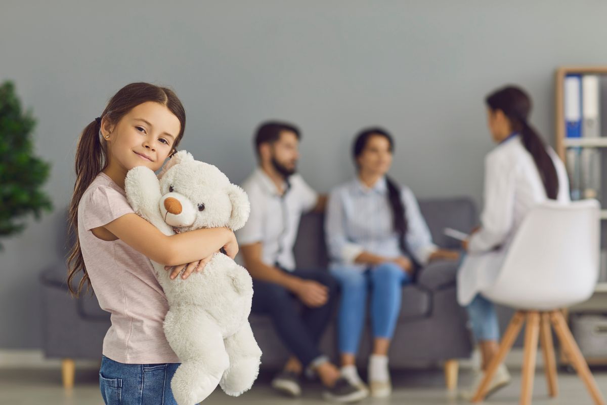 au premier plan une petite fille heureuse fait un calin à un doudou nounours peluche, et en arrière plan le papa et la maman discutent rassurés avec le docteur