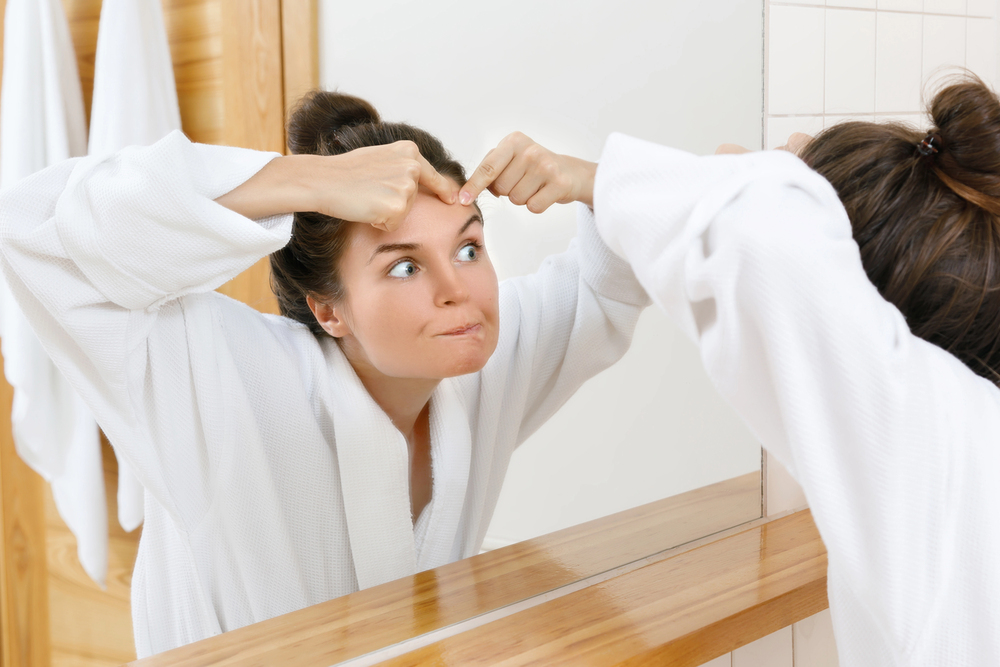 Femme en peignoir qui tente de percer un bouton devant le miroir. Jeune femme pressant un bouton sur son front devant le miroir, geste à éviter en cas d’excès de sébum