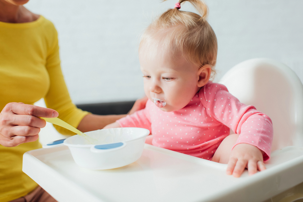 Petite fille bébé enb rose assise dans sa chaise haute en train d’être nourrie à la cuillère par un adulte