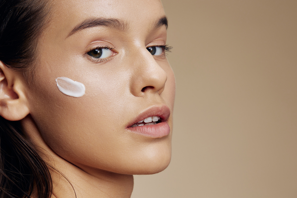 Femme avec une crème sur la joue sur fond beige Femme appliquant une crème visage légère pour peau grasse ou mixte