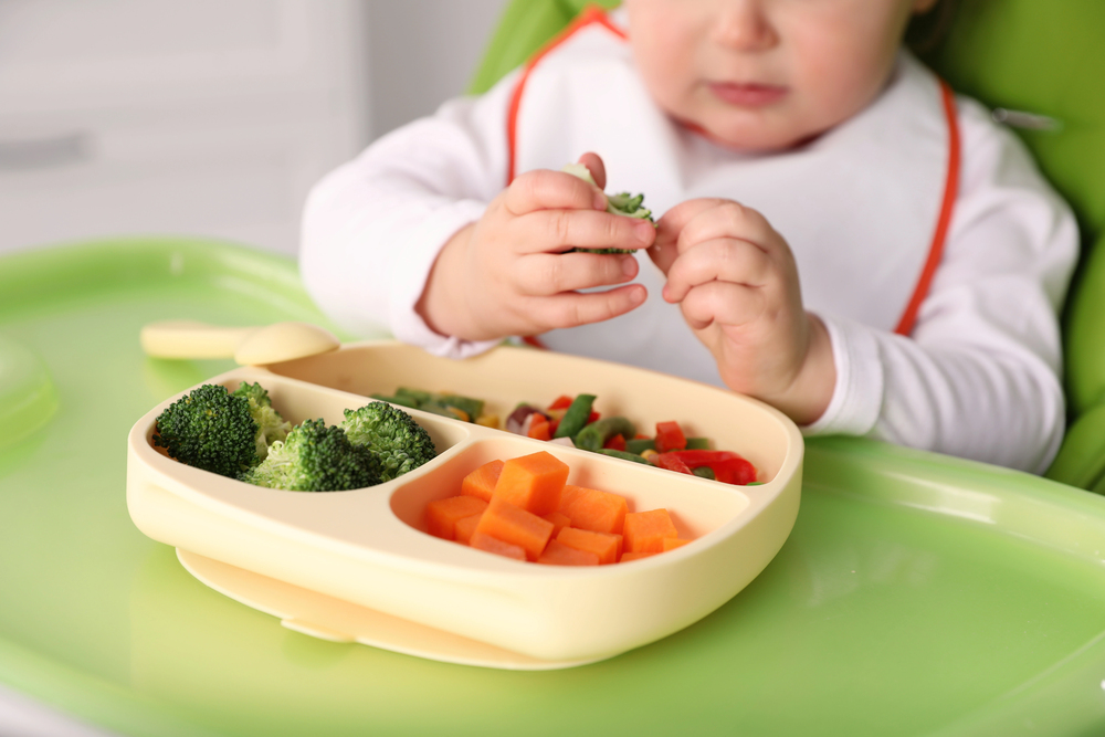 Bébé attrapant un morceau de brocoli dans une assiette compartimentée de légumes vapeur, focus sur les mains