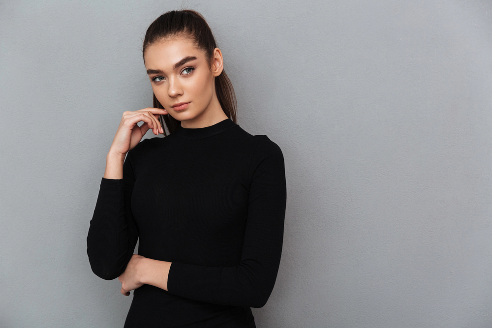 Jeune femme en pull noir ajusté devant un mur gris clair, pose minimaliste et assurée