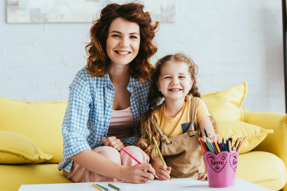Jeune femme nounou souriante avec une petite fille, moment complice à la maison lors d’une garde d’enfants