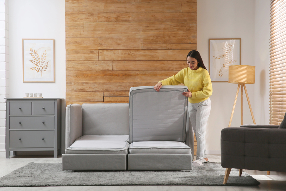 Femme ouvrant un canapé convertible gris dans un salon lumineux et moderne avec décor en bois clair