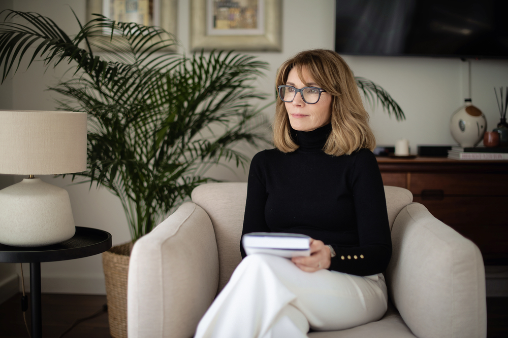 Femme élégante 50ans en pull noir et pantalon blanc dans un salon design, assise avec un carnet, ambiance intellectuelle et cosy