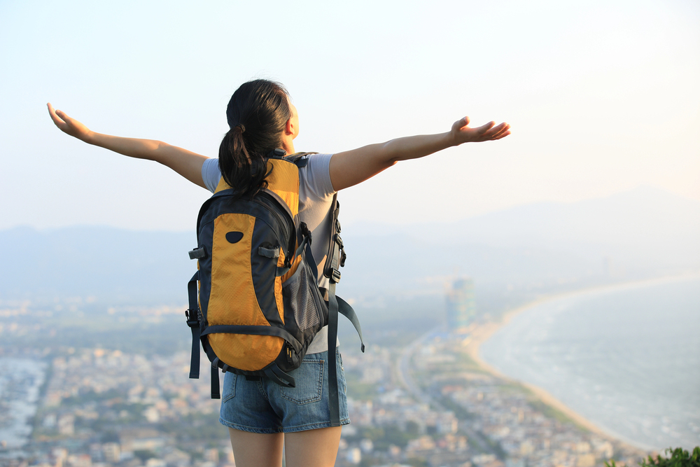 Femme seule les bras ouverts face à la mer, sac à dos, vue en hauteur sur la ville