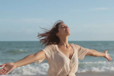 Une femme face à la mer, yeux fermés et bras ouverts, illustrant la liberté et l’équilibre du lâcher prise en amour.