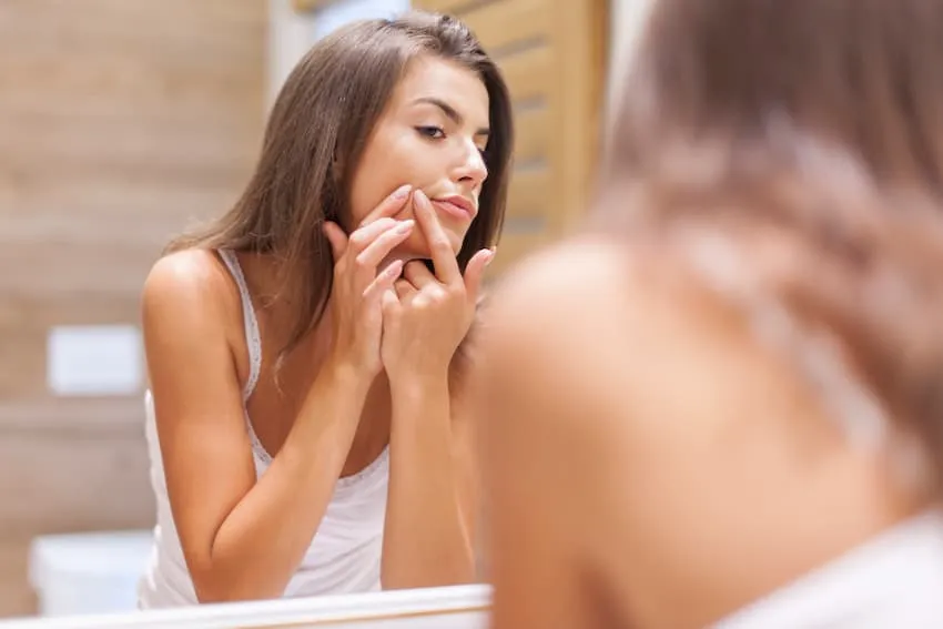 jeune femme adolescente pressant un bouton devant son miroir, geste à éviter pour ne pas aggraver les imperfections.