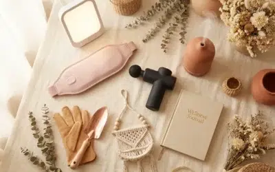 Sélection de cadeaux utiles pour femmes disposés en flat lay élégant : ceinture chauffante, lampe de luminothérapie, pistolet de massage, kit jardinage en cuir naturel, carnet bien-être et diffuseur céramique sur fond lin crème avec fleurs séchées