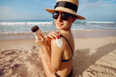 Femme avec chapeau de paille et lunettes de soleil appliquant un écran solaire sur l'épaule à la plage, combo protection solaire complète.
