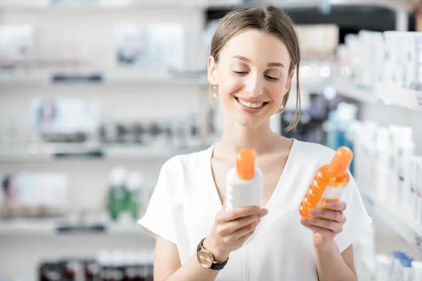 Jeune femme comparant deux flacons de protection solaire en pharmacie, sélection de soin anti-âge et filtres UV.