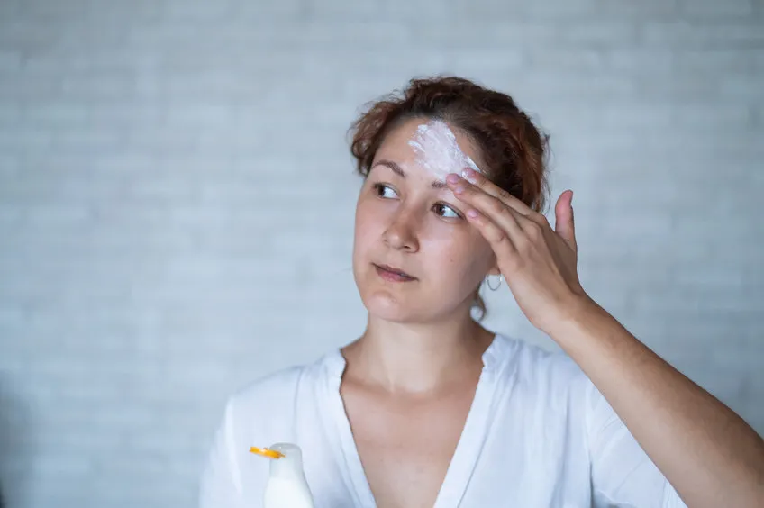 Gros plan d'une femme appliquant une noisette de crème solaire blanche sur le front, prévention des rides et taches solaires.