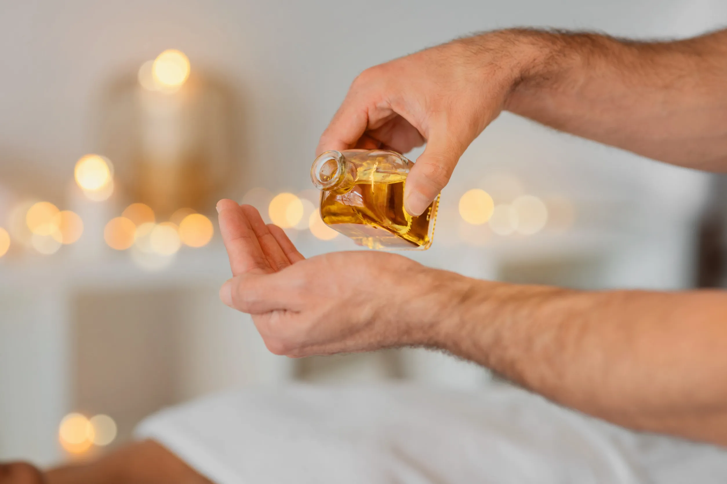 Gros plan d'une main versant de l'huile de massage dorée depuis un flacon en verre dans la paume d'une autre main.