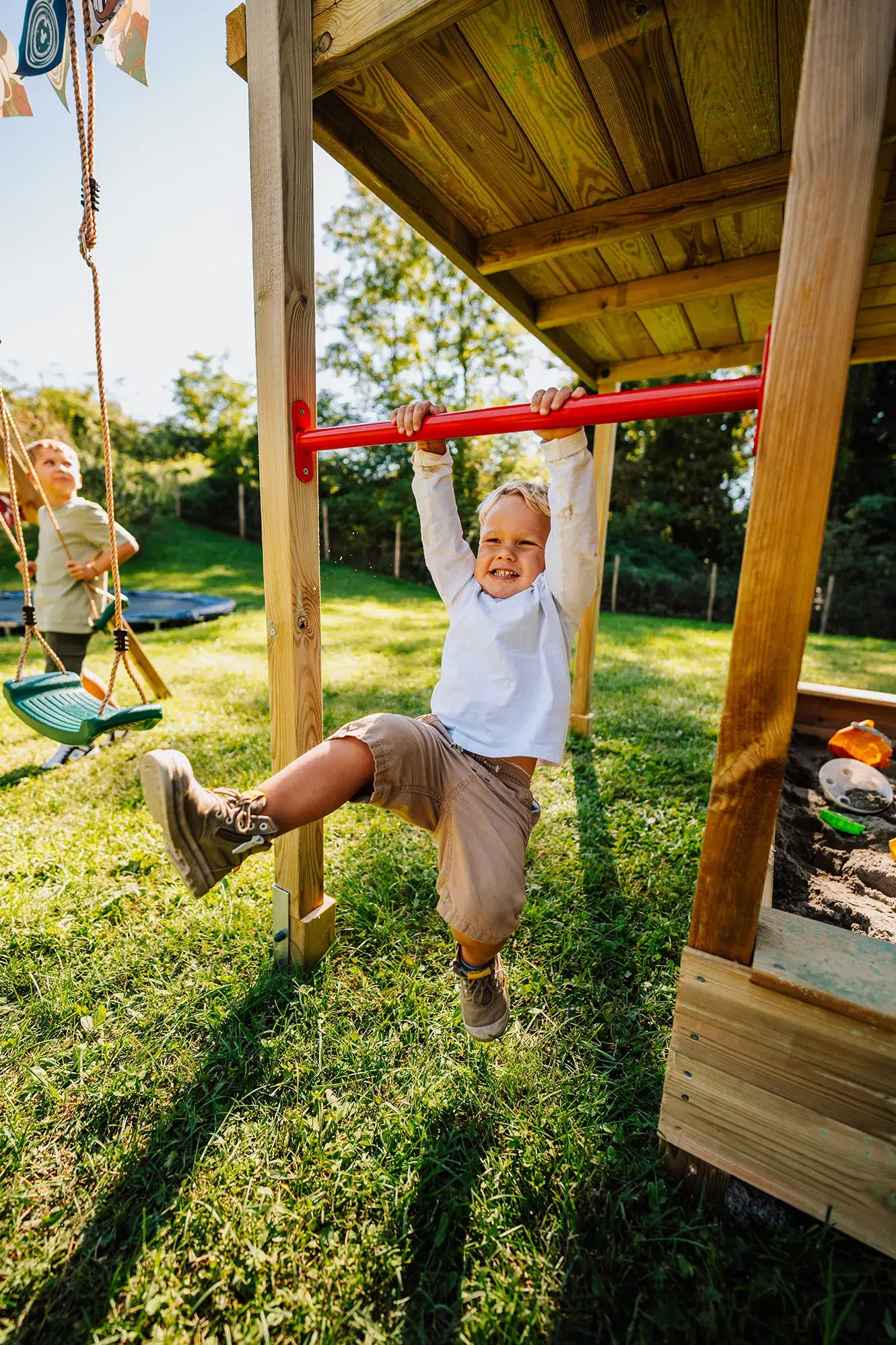 Enfant suspendu à une barre de traction sur un portique en bois, souriant au jardin