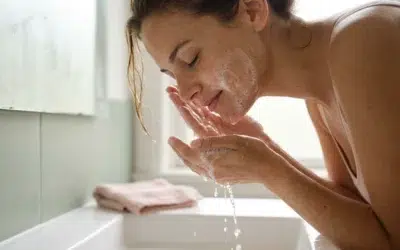 nettoyant-visage-choisir-cover femme à la peau olive rinçant une mousse de nettoyant visage au-dessus d'un lavabo, lumière du matin