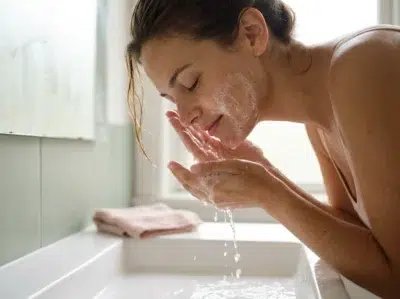 femme à la peau olive rinçant une mousse de nettoyant visage au-dessus d'un lavabo, lumière du matin