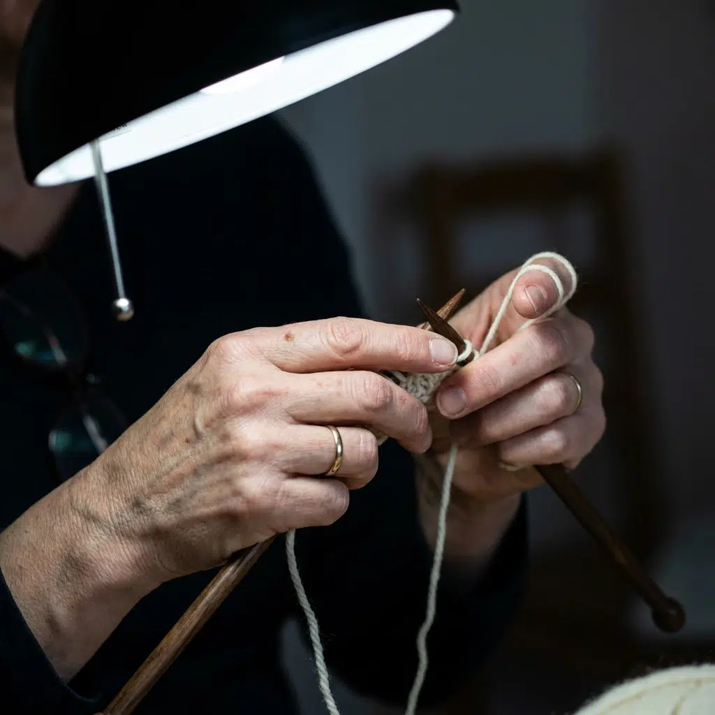 Mains de femme tricotant une laine claire sous une lampe articulée, aiguilles foncées