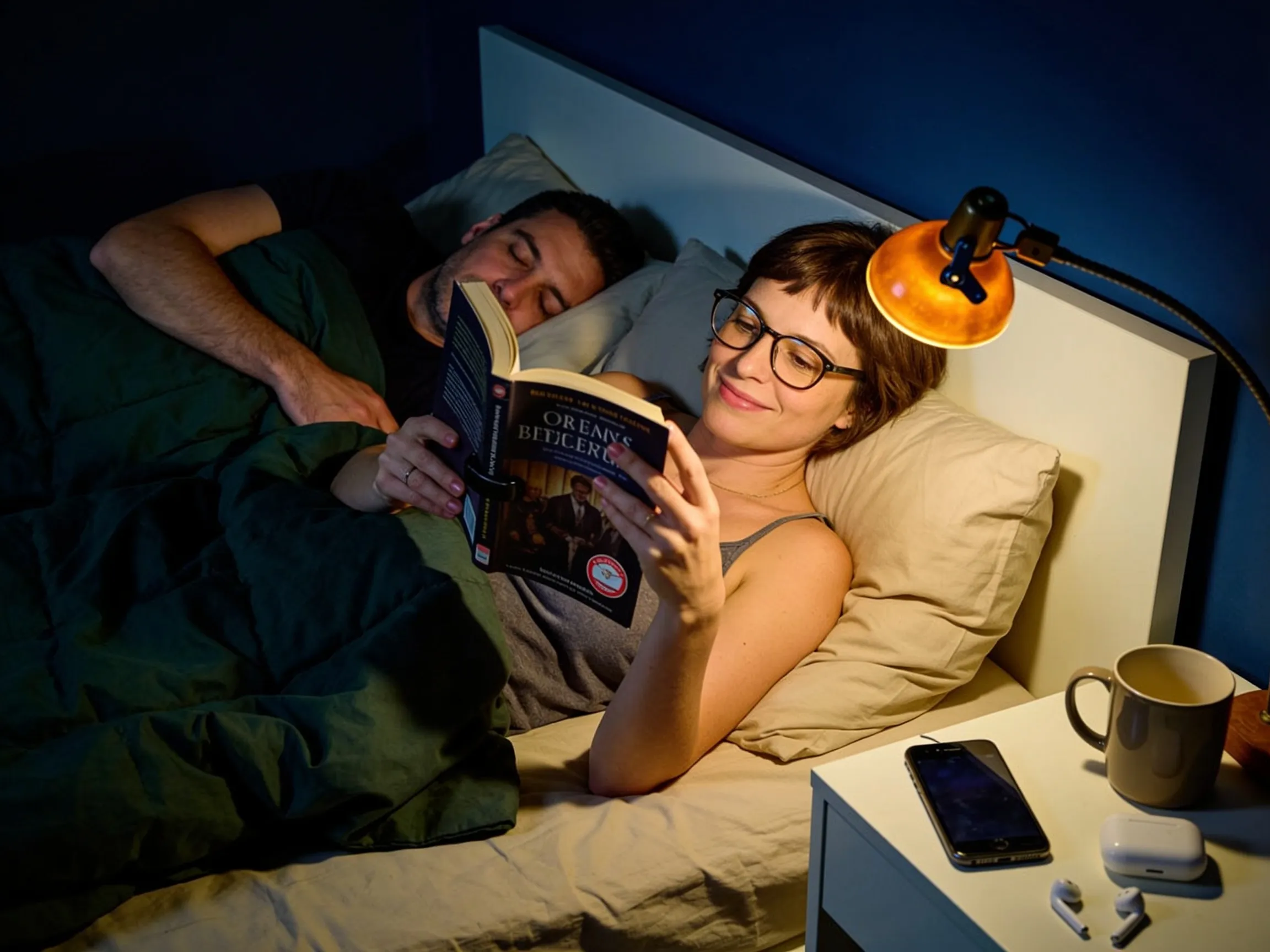 Femme lisant à la lueur d'une petite lampe pendant que sa compagne dort sous sa propre couette dans un grand lit